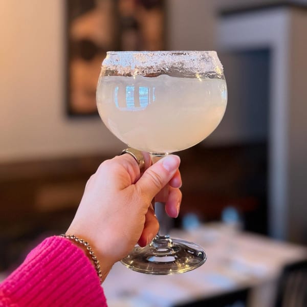 A hand in a pink sweater holds a large, stemmed glass with a salted rim and a light-colored drink.