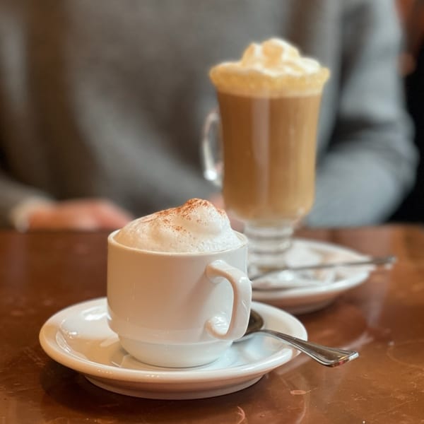 Two frothy coffee drinks, one in a white cup and one in a tall glass, on a wooden table.