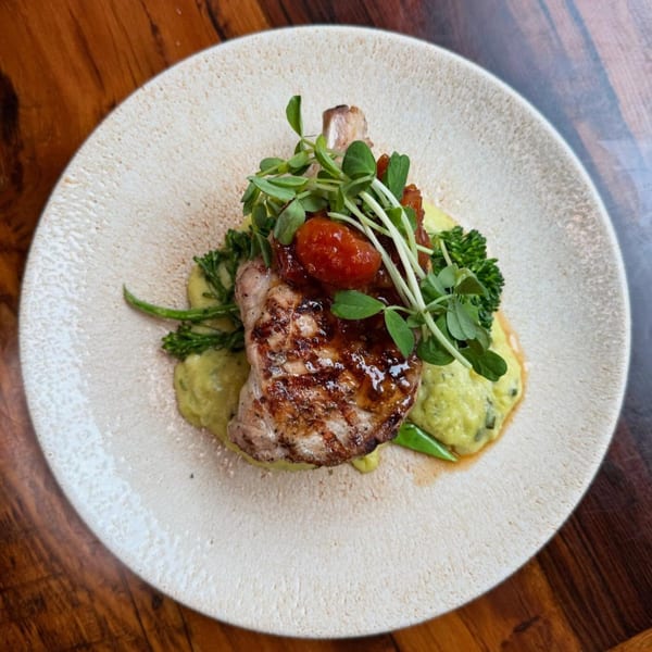 Grilled pork chop with tomato relish, pea shoots, and green puree on a white plate.