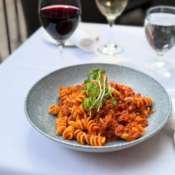 A plate of fusilli pasta with meat sauce and fresh greens, alongside wine and water glasses.