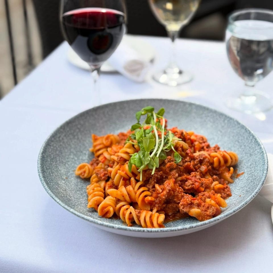 A plate of fusilli pasta with meat sauce and fresh greens, alongside wine and water glasses.