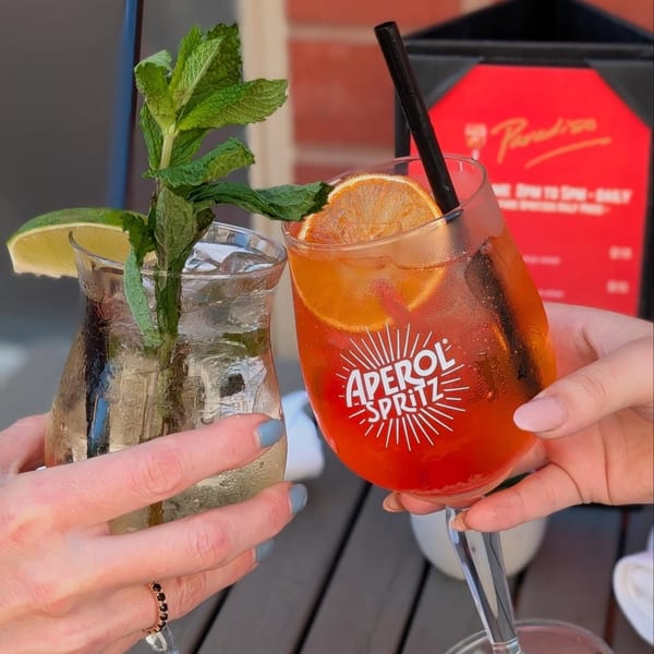 Two hands clinking glasses of Aperol Spritz and a clear cocktail with mint and lime.