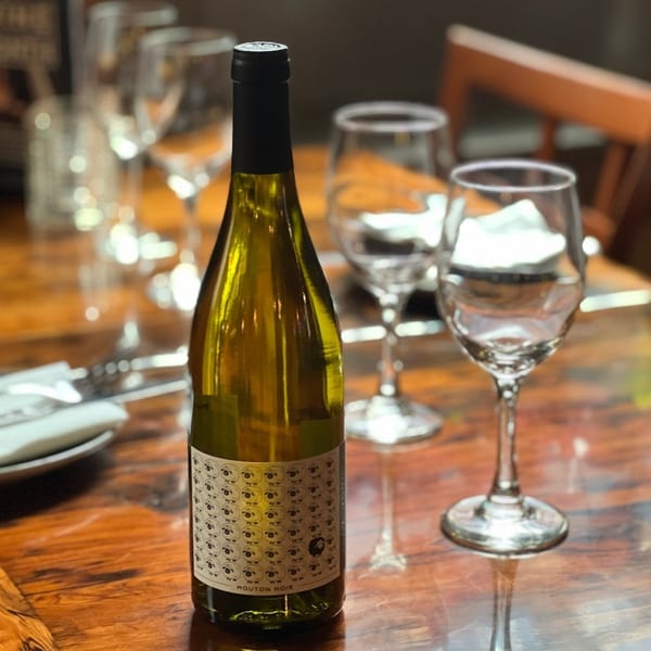 A bottle of Mouton Noir white wine on a wooden table with several empty wine glasses.