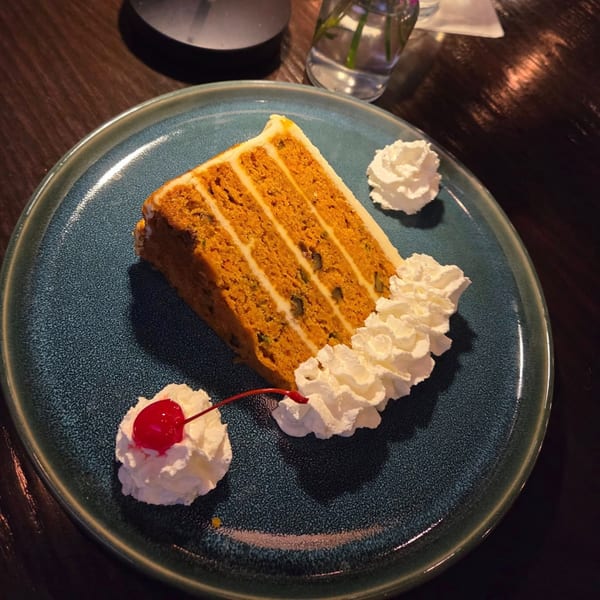 A slice of layered carrot cake with white frosting, whipped cream, and a cherry on a blue plate.