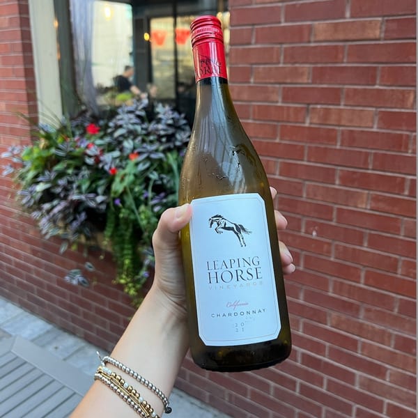 A hand holds a bottle of Leaping Horse Chardonnay wine against a brick wall and green plants.