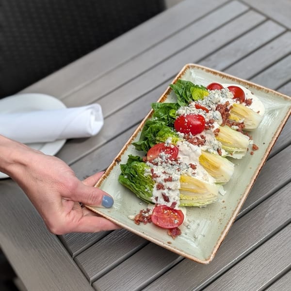 A hand presenting a rectangular plate with a wedge salad topped with tomatoes, bacon, and blue cheese.