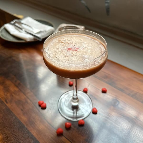 A foamy brown cocktail in a coupe glass, garnished with a red heart, on a wooden table.
